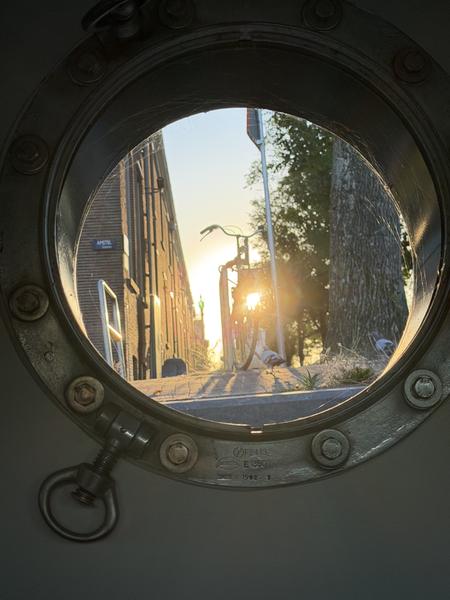 This photo captures a serene street scene in Amstel, Weesperbuurt, Amsterdam, viewed through the circular frame of a porthole